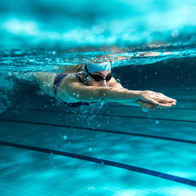 Une femme qui nage en piscine, en mouvement, vue sous l'eau. Elle porte des lunettes et un bonnet.